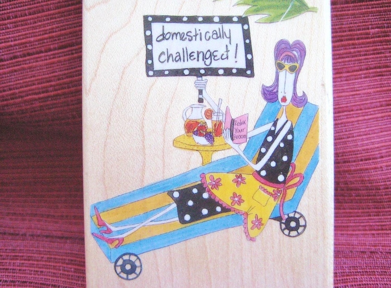 Op de afbeelding: Houten blok met een cartoon illustratie van een vrouw die ontspant op een ligstoel. Ze leest een boek met de tekst "Relax Your Broom" en heeft een drankje op een tafeltje. Het bord boven haar zegt "domestically challenged!"