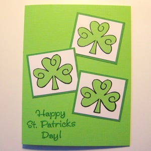 May include: A green St. Patrick's Day greeting card with three shamrock illustrations. Each shamrock is outlined in black and set on a white square with a green border. The words "Happy St. Patrick's Day!" are printed in green.