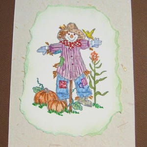 May include: Hand-drawn illustration of a scarecrow with a straw hat, red and white checkered scarf, and patched blue jeans. A yellow bird perches on its arm. Two orange pumpkins sit at the base. The artwork is on textured paper with a green border.