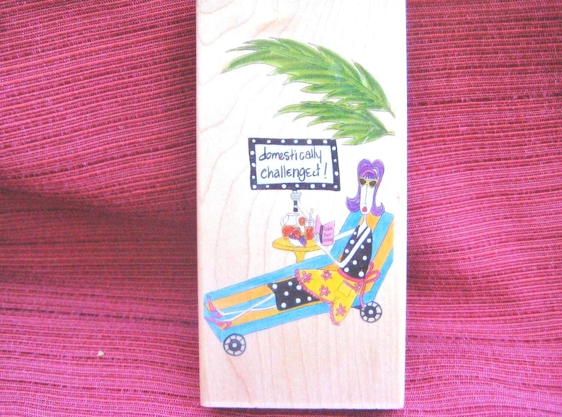 Op de afbeelding: Houten blok met een grappige illustratie van een persoon die ontspant op een ligstoel. De persoon draagt een zonnebril en leest een boek, met een drankje en fruit op een bijzettafeltje. De tekst "domestically challenged!" is afgebeeld.