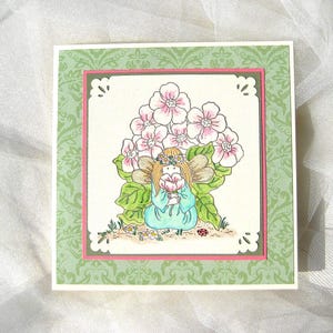 May include: A square greeting card featuring a whimsical illustration of a fairy holding a pink flower, surrounded by large pink flowers and green leaves. The card has a green patterned border with a pink inner border and decorative cutouts.