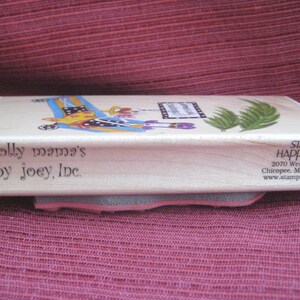 Op de afbeelding: Een houten stempel met de tekst "dolly mama's by joey, Inc." en een kleurrijke illustratie. De stempel heeft een grijze en roze rubberen basis. Op de zijkant van de stempel staat "STAMPS HAPPEN, INC."