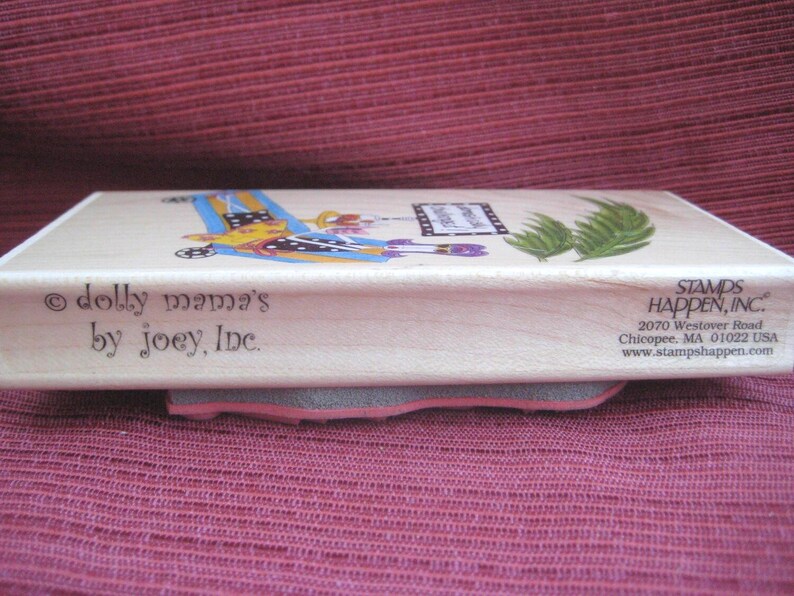 Op de afbeelding: Een houten stempel met de tekst "dolly mama's by joey, Inc." en een kleurrijke illustratie. De stempel heeft een grijze en roze rubberen basis. Op de zijkant van de stempel staat "STAMPS HAPPEN, INC."
