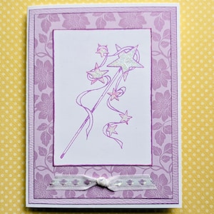 May include: A handmade greeting card with a purple floral design. The card features a central white rectangle with a purple illustration of a star wand with ribbons and stars. A silver ribbon with a bow is at the bottom.