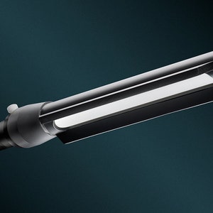 May include: A black and silver handheld LED light with a long, rectangular shape and a round end. The light is turned on and illuminating the surrounding area.