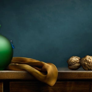 May include: A still life composition featuring a green glass vase with blue accents, a golden cloth, and three walnuts. The vase is round with a narrow neck. The walnuts are textured and golden brown.