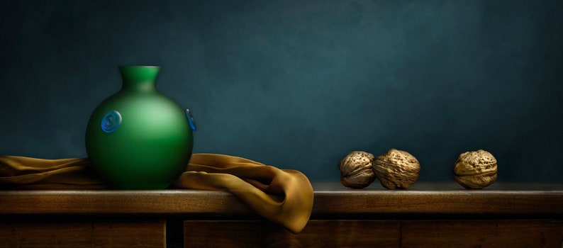 May include: A still life composition featuring a green glass vase with blue accents, a golden cloth, and three walnuts. The vase is round with a narrow neck. The walnuts are textured and golden brown.