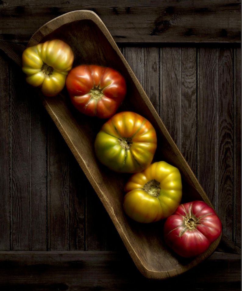 May include: A wooden trough filled with heirloom tomatoes. The tomatoes range in color from yellow-green to deep red. The tomatoes are arranged in a row inside the wooden trough, which sits on a dark wooden surface.