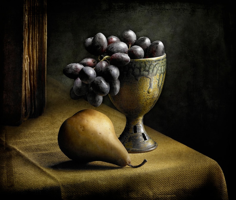 May include: A still life composition featuring a pear and a bunch of dark grapes spilling from a decorative, aged ceramic chalice. The chalice has a textured, dripped glaze. The fruit rests on a golden-brown cloth, with a dark background.