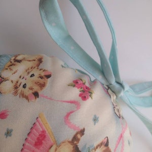 May include: A light blue drawstring bag with white polka dots. The bag is made of fabric and has a print of kittens wearing pink tutus and holding pink ribbons. The bag is tied with a light blue ribbon.