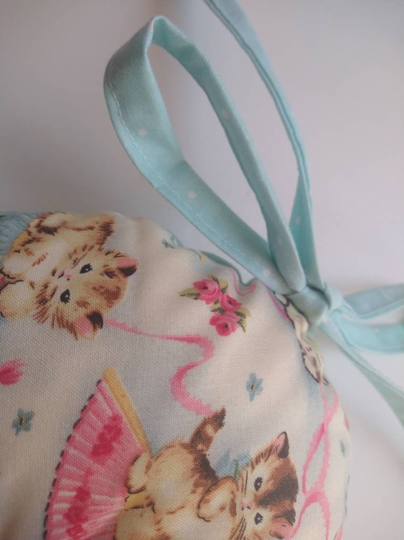 May include: A light blue drawstring bag with white polka dots. The bag is made of fabric and has a print of kittens wearing pink tutus and holding pink ribbons. The bag is tied with a light blue ribbon.