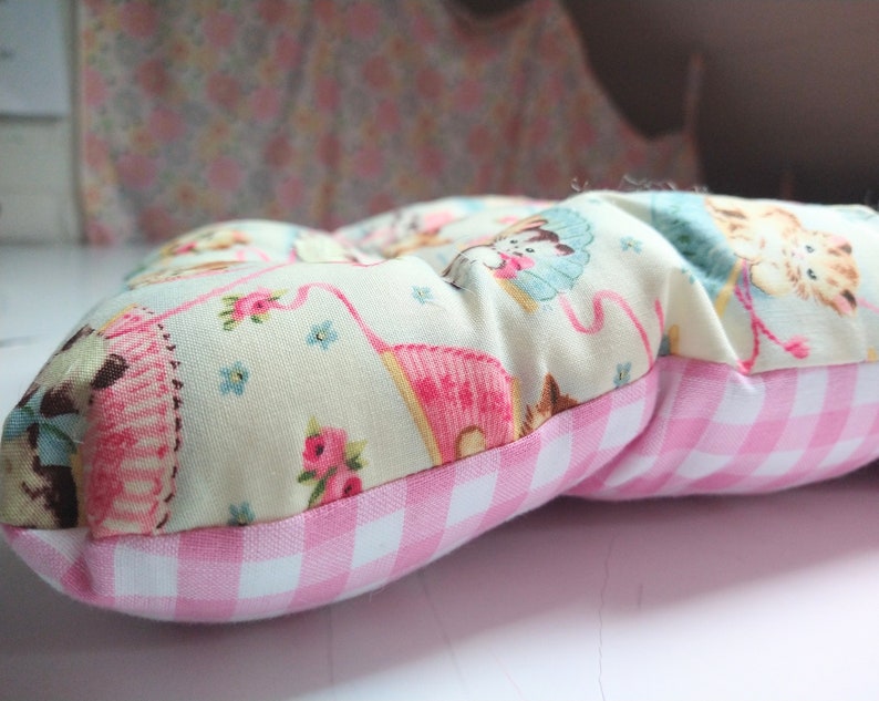 May include: A pink and white gingham cushion with a cream fabric top featuring a repeating pattern of kittens and flowers.