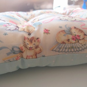 May include: A light blue and white cushion with a floral pattern featuring kittens. The cushion has a light blue border and is filled with stuffing.