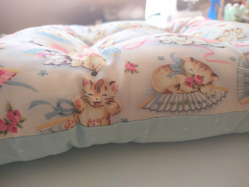 May include: A light blue and white cushion with a floral pattern featuring kittens. The cushion has a light blue border and is filled with stuffing.