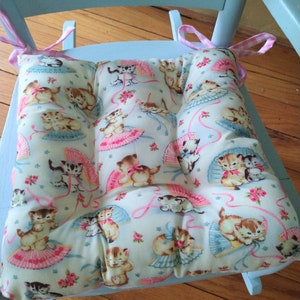 May include: A light blue rocking chair with a white and pink floral cushion featuring a pattern of kittens wearing pink bows and holding fans. The cushion has a light blue background with pink ribbons and white stars.