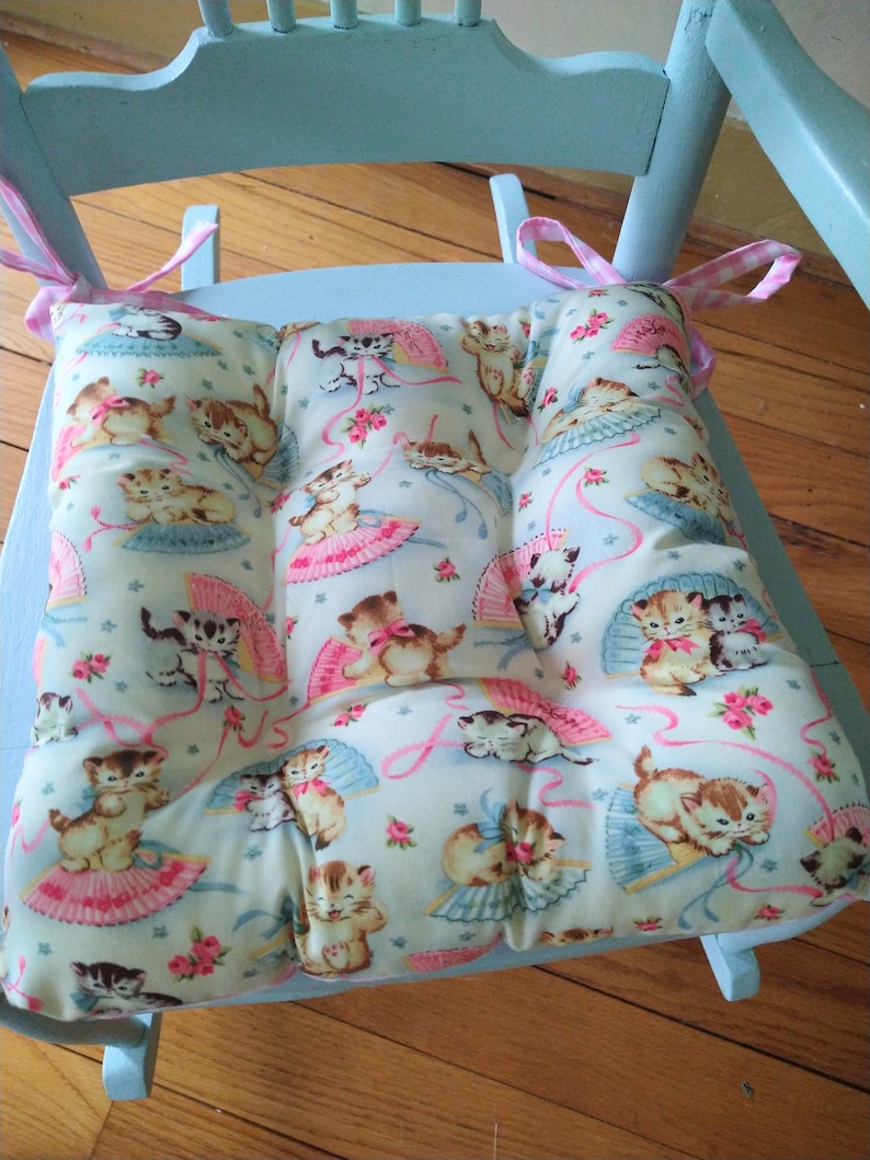 May include: A light blue rocking chair with a white and pink floral cushion featuring a pattern of kittens wearing pink bows and holding fans. The cushion has a light blue background with pink ribbons and white stars.