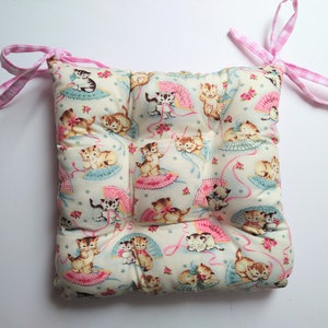 May include: A light blue and white chair cushion with a pattern of kittens holding fans. The cushion has pink and white gingham ties.