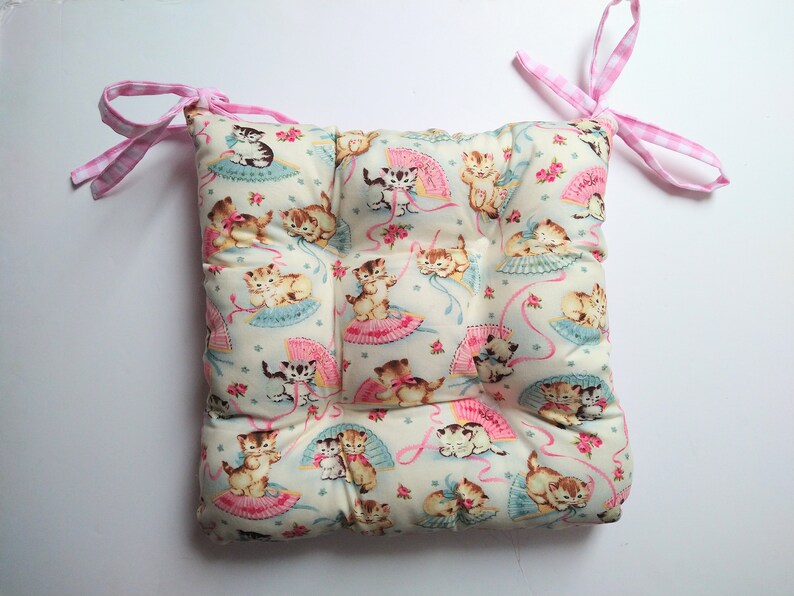 May include: A light blue and white chair cushion with a pattern of kittens holding fans. The cushion has pink and white gingham ties.
