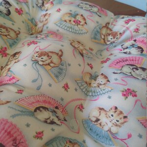 May include: A white cushion with a light blue background and a pattern of pink and blue fans with kittens holding them. The kittens are wearing pink bows and are surrounded by pink and blue ribbons.