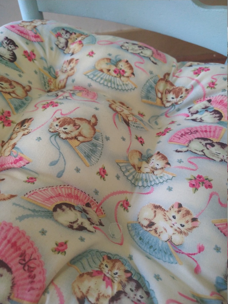 May include: A white cushion with a light blue background and a pattern of pink and blue fans with kittens holding them. The kittens are wearing pink bows and are surrounded by pink and blue ribbons.