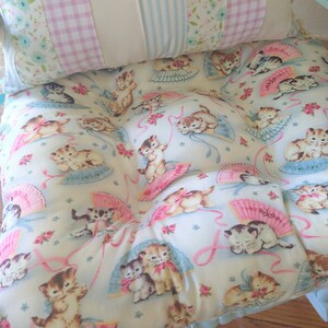 May include: A light blue and white cushion with a floral pattern and a repeating image of kittens holding fans. The cushion is made of fabric and has a ruffled edge.