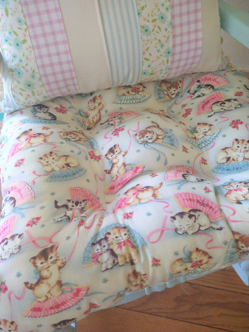 May include: A light blue and white cushion with a floral pattern and a repeating image of kittens holding fans. The cushion is made of fabric and has a ruffled edge.