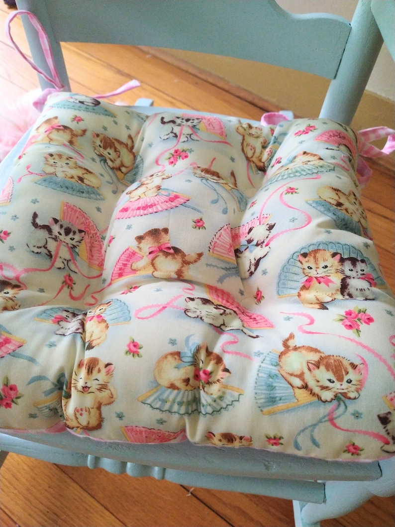 May include: A light blue chair with a pink and white patterned cushion featuring a repeating design of kittens with pink bows and fans. The cushion has pink ties.