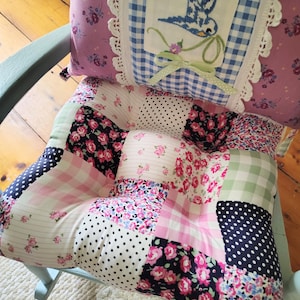 May include: A patchwork seat cushion and backrest set featuring various floral, polka dot, and gingham patterns in pink, white, black, and green. The backrest includes a central blue bird embroidery on a white square with lace trim.