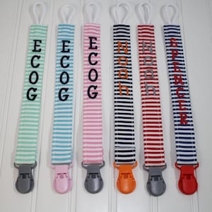 May include: Five striped fabric pacifier clips with different colored clips and personalized names:  'Ecog', 'Ecog', 'Ecog', 'Noah', 'Noah', and 'Spencer'.