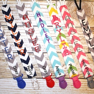 May include: Eight personalized pacifier clips with chevron patterns in various colors, including gray, navy, aqua, pastel, and hot pink. Each clip features a different name, such as Carter, Miller, Anslee, Hadley, Brylie, Anslee, Emilio.