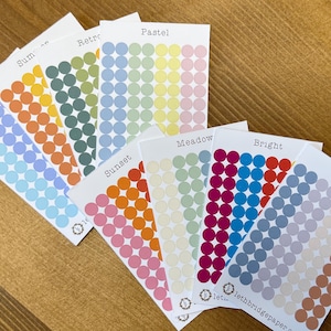 Puede incluir: Una colección de pegatinas de puntos de colores dispuestas en tarjetas blancas. Las tarjetas están etiquetadas con temas de color como "Summer", "Retro" y "Pastel". Las pegatinas son de varios tonos de naranja, verde, azul y rosa. El fondo es una superficie de madera.