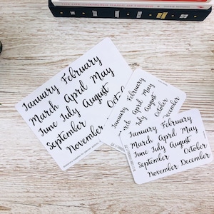 May include: Three white cards with black handwritten text listing the months of the year. The cards are on a white wooden surface.