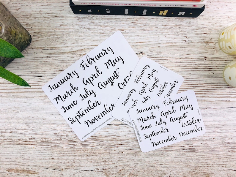 May include: Three white cards with black handwritten text listing the months of the year. The cards are on a white wooden surface.