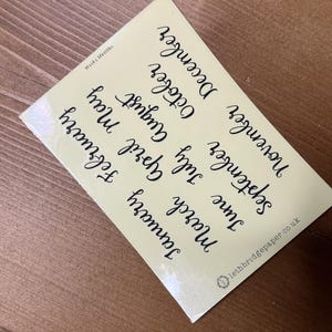 May include: A light yellow sticker featuring the months of the year in black cursive lettering. The text "Weeks Monks" is at the top left, and "lethbridgepaper.co.uk" is at the bottom right. The sticker is on a wooden surface.