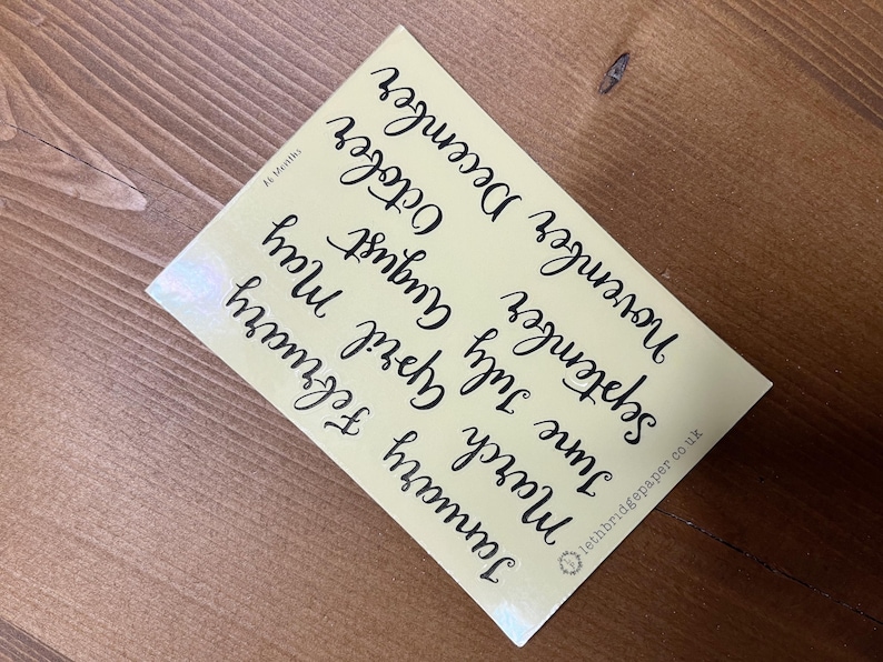 May include: A pale yellow card featuring the months of the year in black cursive script. The card is angled on a wooden surface. The text "lethbridgepaper co uk" is visible.