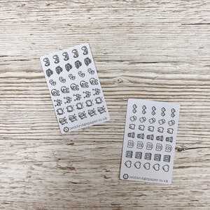 May include: Two sheets of white sticker paper with black outlines of various icons. The first sheet has icons of skulls, crossbones, and other spooky designs. The second sheet has icons of glasses, teeth, coffee cups, cameras, and hearts. Both sheets have the text "lethbridgepaper.co.uk" at the bottom.