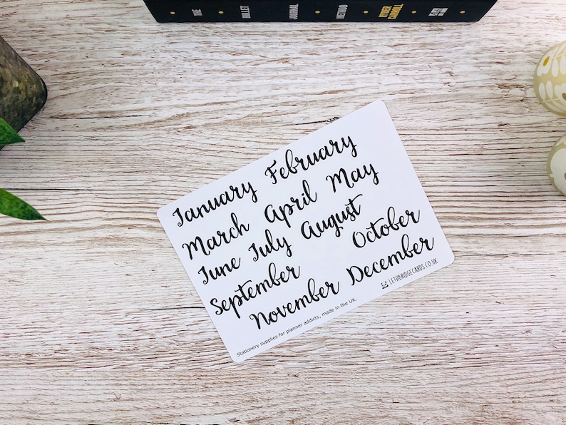 May include: White card with black handwritten text listing the months of the year: January, February, March, April, May, June, July, August, September, October, November, December. The text "Stationery supplies for planner addicts, made in the UK" is printed at the bottom of the card.