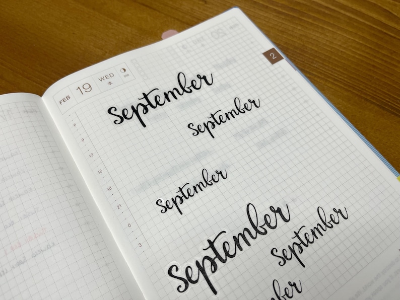 May include: An open notebook with grid paper, displaying the word "September" in a cursive font several times. The date "FEB 19 WED" is visible. The notebook is on a wooden surface.