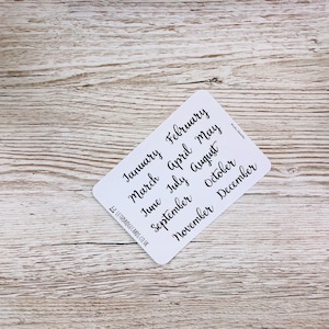 May include: A white card with the months of the year written in black cursive script. The months are: January, February, March, April, May, June, July, August, September, October, November, December.