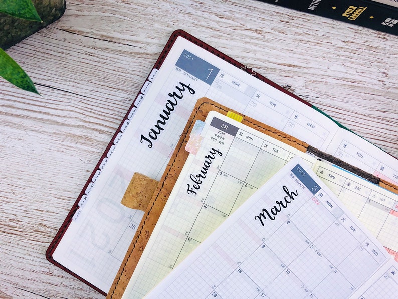 May include: A close-up of three open planners with monthly calendars for January, February, and March. The planners are brown leather with cork accents and have Japanese text.