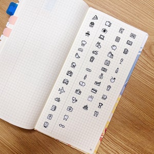 May include: A page from a notebook with a grid pattern. The page is filled with black and white icons representing various activities, objects, and concepts. Some of the icons include a pizza slice, a camera, a house, a plane, a coffee cup, a tooth, and a pair of glasses.
