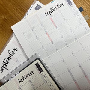 Può includere: Diversi planner aperti con il mese di settembre visualizzato. La parola "Settembre" è scritta in corsivo sulle copertine. Le pagine mostrano griglie di calendario con date e del testo giapponese. I planner sono su una superficie di legno.