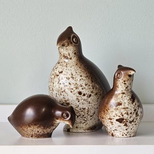 May include: A set of three ceramic bird figurines with a speckled brown and cream glaze. The largest bird stands upright, while the two smaller birds are positioned lower, one with a rounded body and the other with a more compact, upright shape.