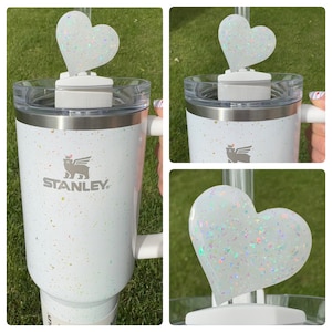 May include: White stainless steel travel mug with a white glitter finish. The mug has a black logo that says "STANLEY". A white glitter heart-shaped straw topper is on top of the mug.