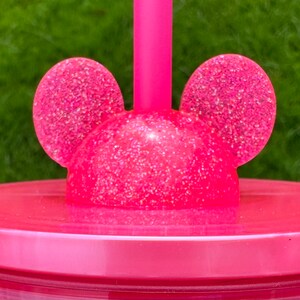 May include: Pink plastic cup with a glitter finish and a lid featuring two pink glitter mouse ears. The lid has a hole for a straw.