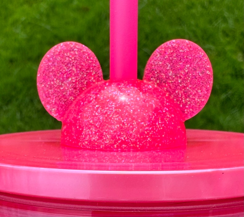 May include: Pink plastic cup with a glitter finish and a lid featuring two pink glitter mouse ears. The lid has a hole for a straw.