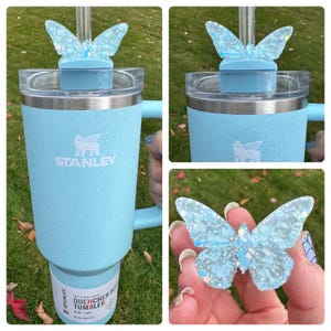 May include: A light blue stainless steel tumbler with a black Stanley logo and a butterfly-shaped lid. The lid is clear and has a blue butterfly design with glitter accents. The tumbler is labeled "The Flowstate 40oz Quencher Tumbler".