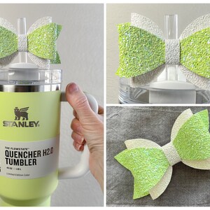 May include: A green and white glitter bow with a white center. The bow is attached to a clear plastic lid. The lid is on top of a green Stanley tumbler with the text "The Flowstate Quencher H2.0 Tumbler 40 oz | 1.18 L Limited Edition Color" printed on the side.