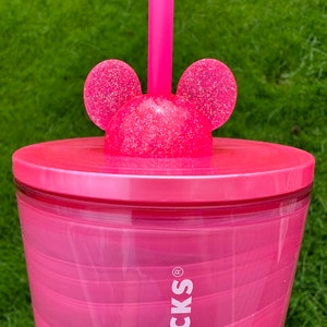 May include: Pink reusable tumbler with a swirl design and a pink lid with glitter and Mickey Mouse ears. The tumbler has the word "BUCKS" printed on the side.
