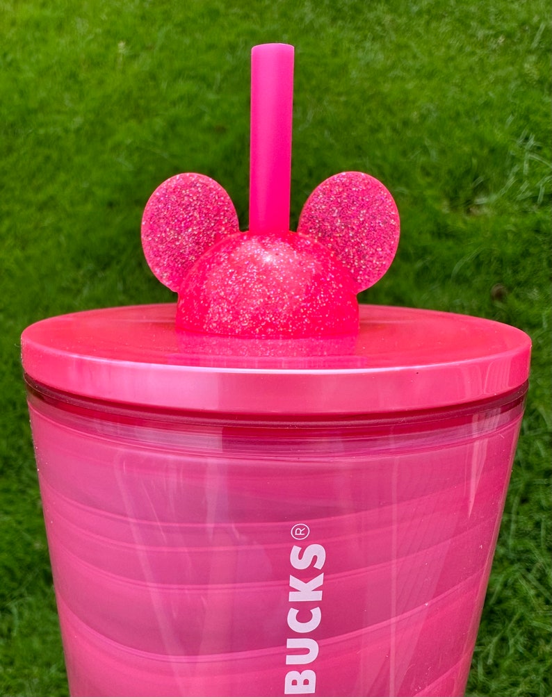 May include: Pink reusable tumbler with a swirl design and a pink lid with glitter and Mickey Mouse ears. The tumbler has the word "BUCKS" printed on the side.